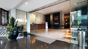A modern hotel lobby with a reception desk, a large potted plant, and a clear view into the kitchen area. - Hotel Nyland Cipaganti (Bandung)