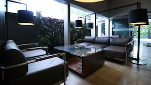 A modern living room with a glass coffee table, leather sofa, and chairs. There are large windows with plants outside. - Hotel Nyland Cipaganti (Bandung)