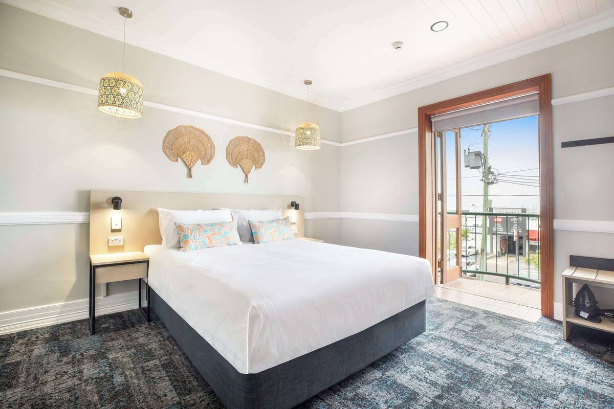 A hotel room with a large bed, two bedside lamps, a small table, a balcony with a view, and decorative wall art.