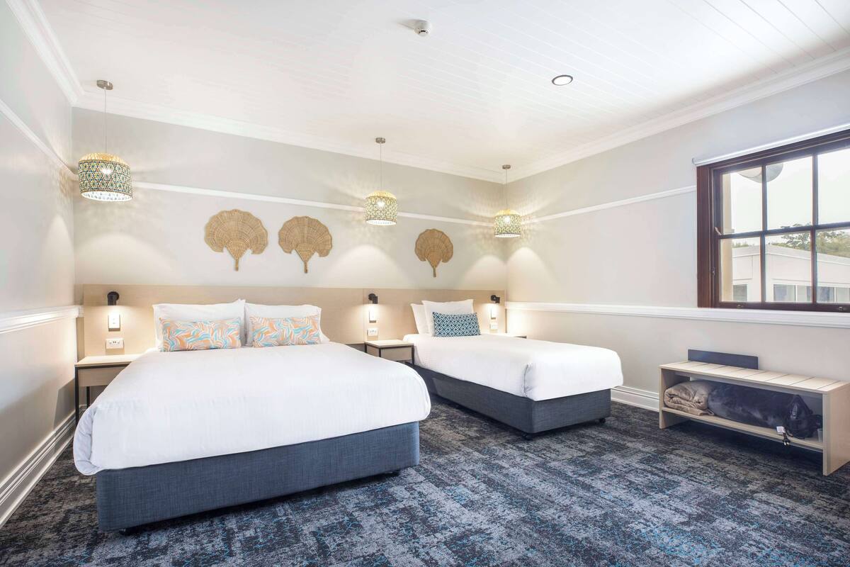 A hotel room with two beds, a small desk, a bench, and a large window.