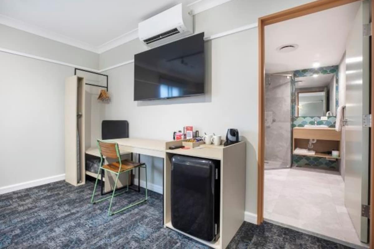 A compact hotel room with a desk, chair, and a small refrigerator. A TV is mounted on the wall, and a bathroom is visible through an open door.