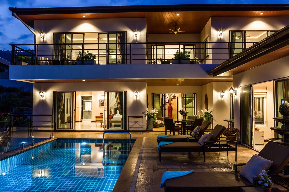 A modern two-story house with a swimming pool, outdoor seating area, and a balcony with plants.