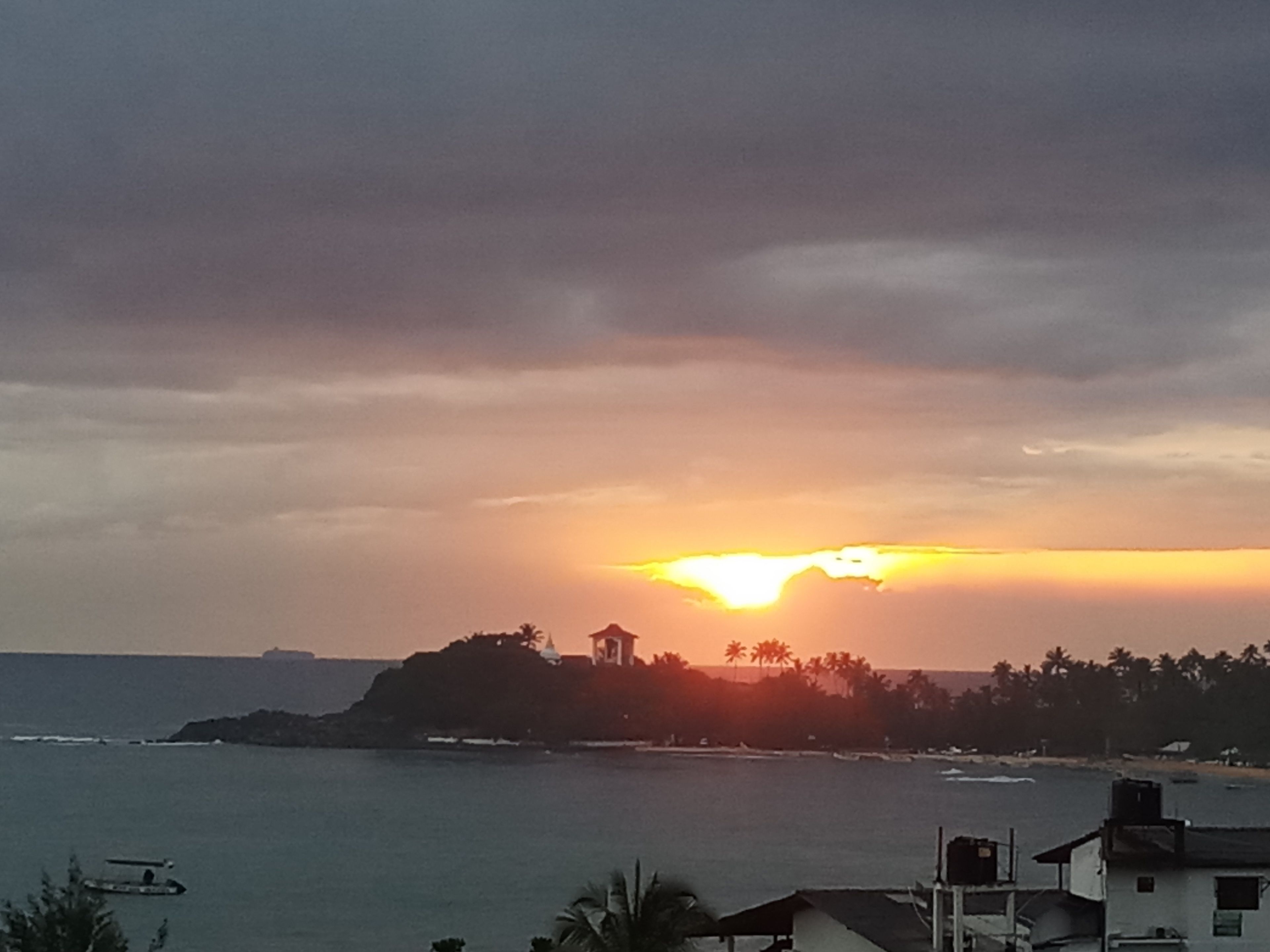 A coastal sunset view with a small island, palm trees, and a boat in the water.