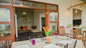 A dining area with a table set for four, a vase with a plant, and a view into a living room with a sofa and cushions. - Sandaya's Place (Unawatuna)