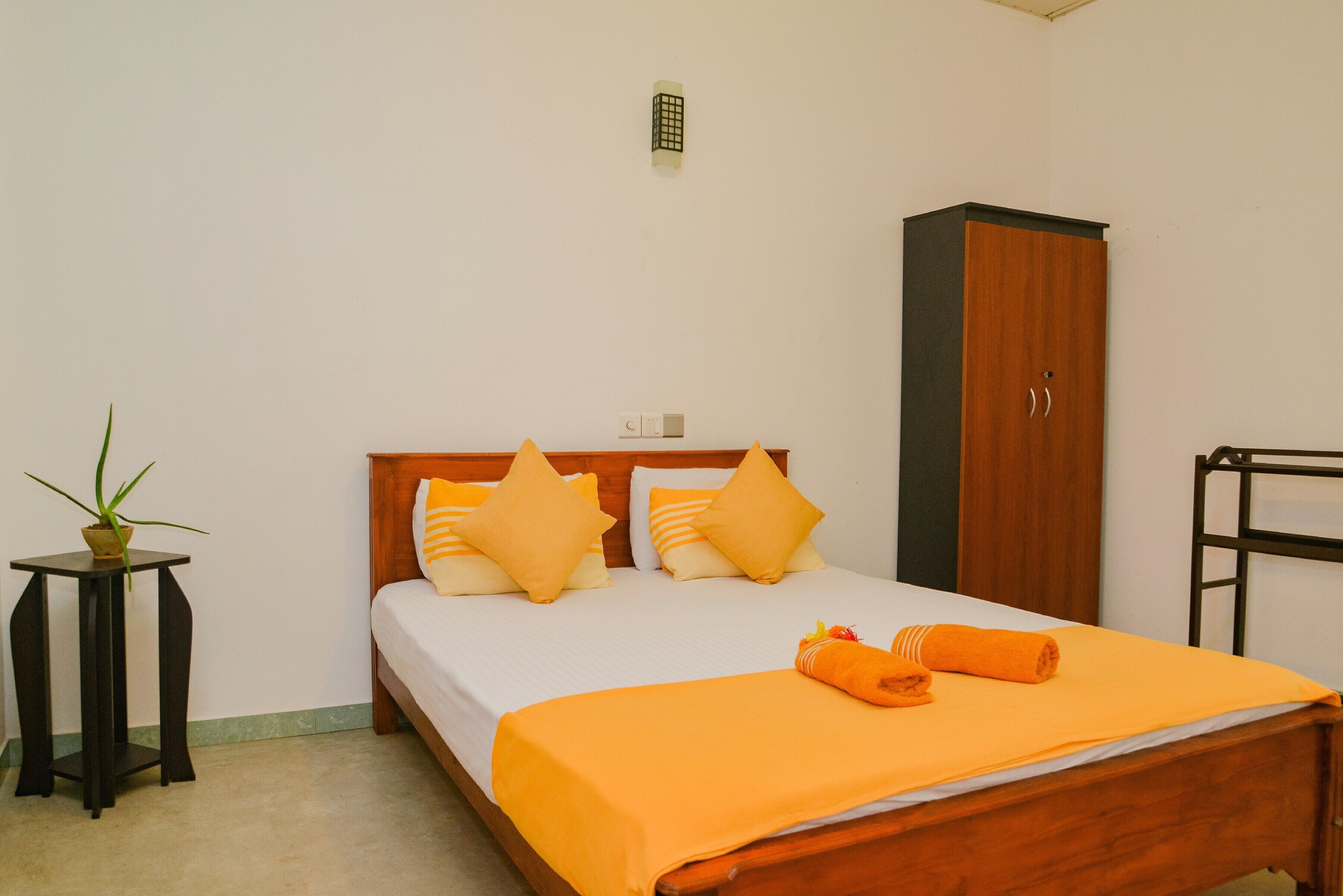 Een opgemaakt bed met een oranje sprei en kussens, een klein tafeltje met een plant en een kast in een eenvoudige kamer.