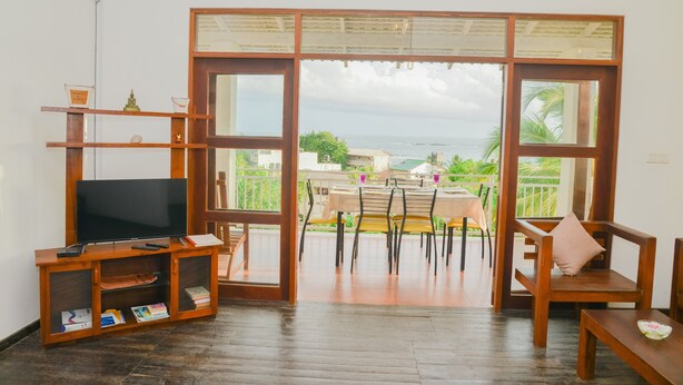A room with a television on a wooden stand, a table with chairs, and a view of the sea through large glass doors. - Sandaya's Place (Unawatuna)