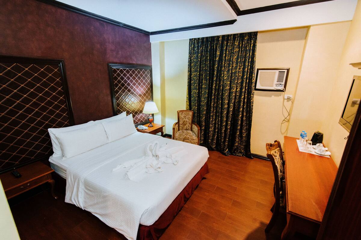 A hotel room with a large bed, a chair, a table, and a wall-mounted air conditioner.