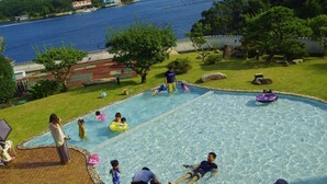 En swimmingpool där folk badar och njuter av vattnet, omgiven av en gräsyta och träd, med utsikt över en sjö eller ett annat vattendrag och byggnader i bakgrunden.