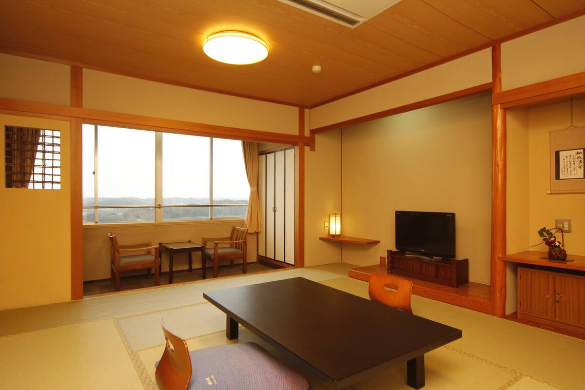 Quarto tradicional japonês com piso de tatame, uma mesa baixa, cadeiras, televisão e vista pela janela grande.