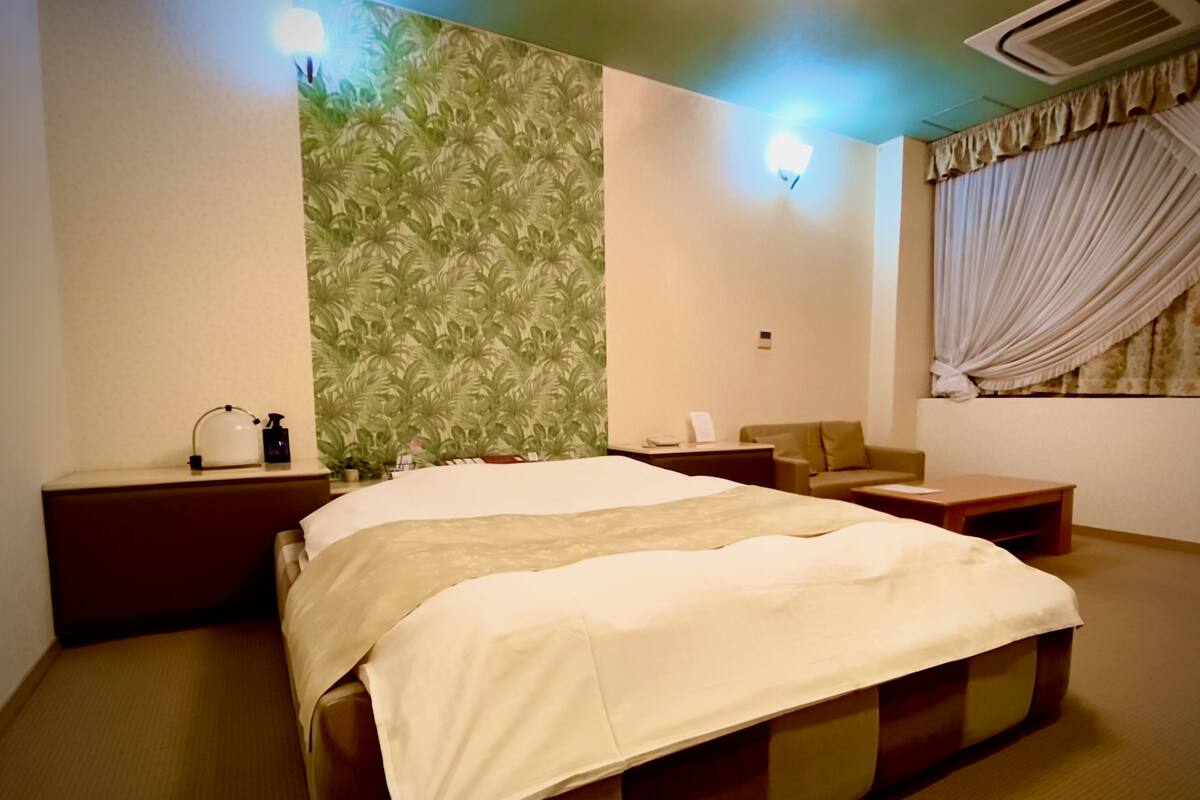 Una habitación de hotel con cama, mesita de noche, silla, una pequeña mesa y ventana con cortinas.