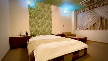 A hotel room with a bed, a nightstand, a chair, a small table, and a window with curtains.