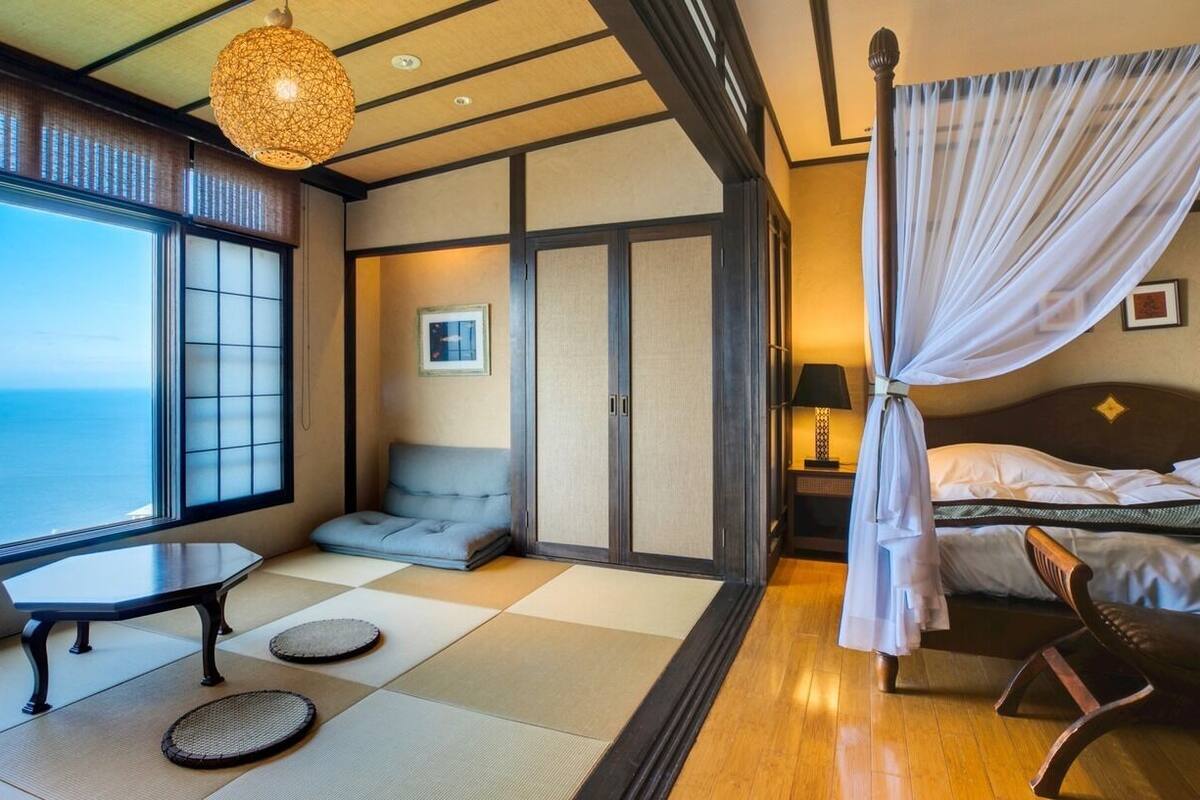 A traditional Japanese-style room with tatami flooring, a low table, a cushioned seating area, a bed with a canopy, and a view of the sea.