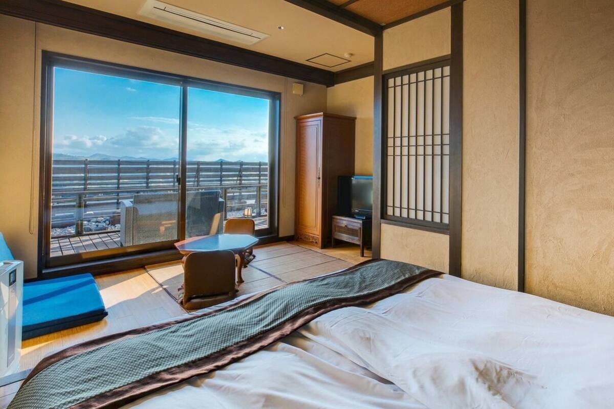 A hotel room with a large sliding glass door, a bed, a small table, a chair, and a television.