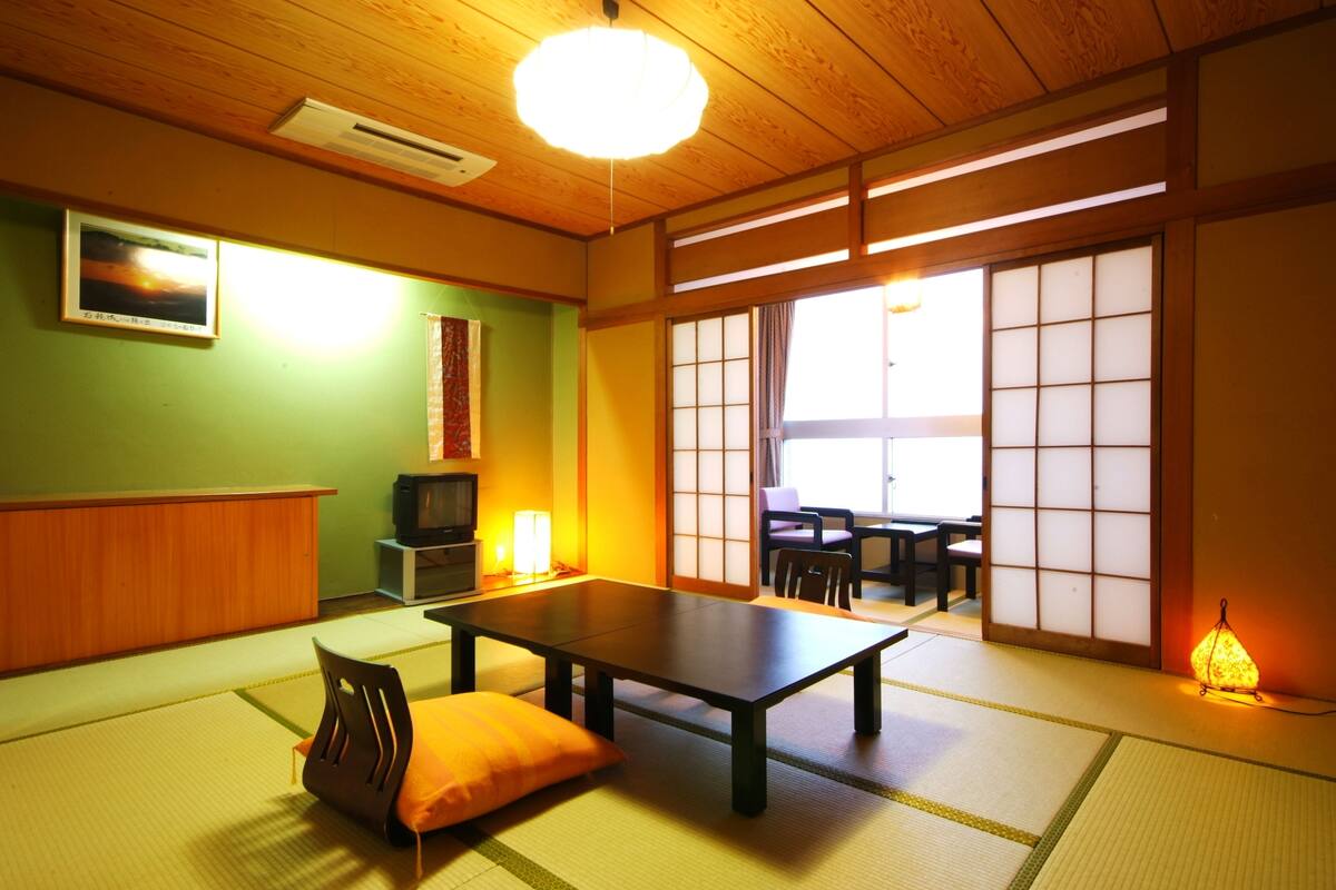 A traditional Japanese room with tatami flooring, a low table, a cushion, a television, and sliding doors leading to a dining area.