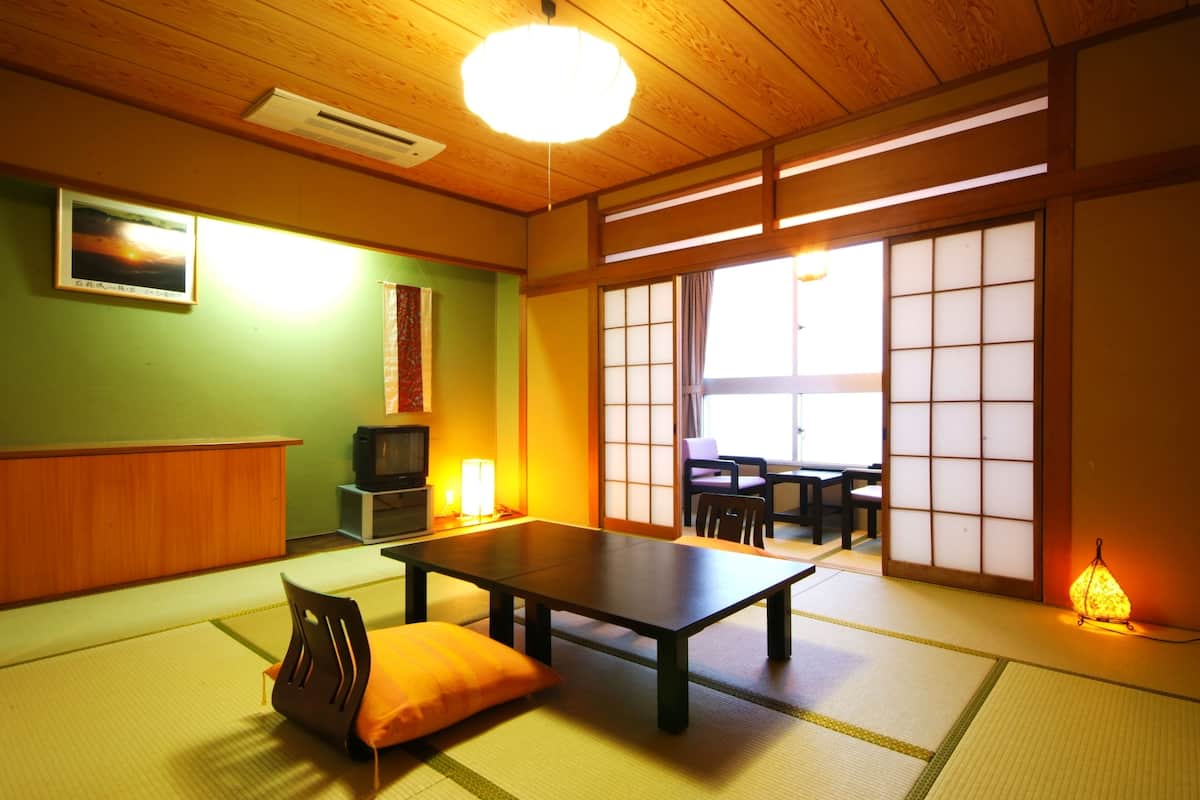A traditional Japanese room with tatami flooring, a low table, a cushion, a television, and sliding doors leading to a dining area.