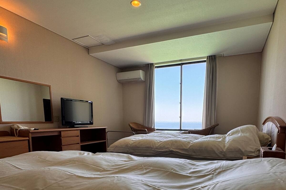 A hotel room with a bed, a desk with a television, a chair, and a large window.