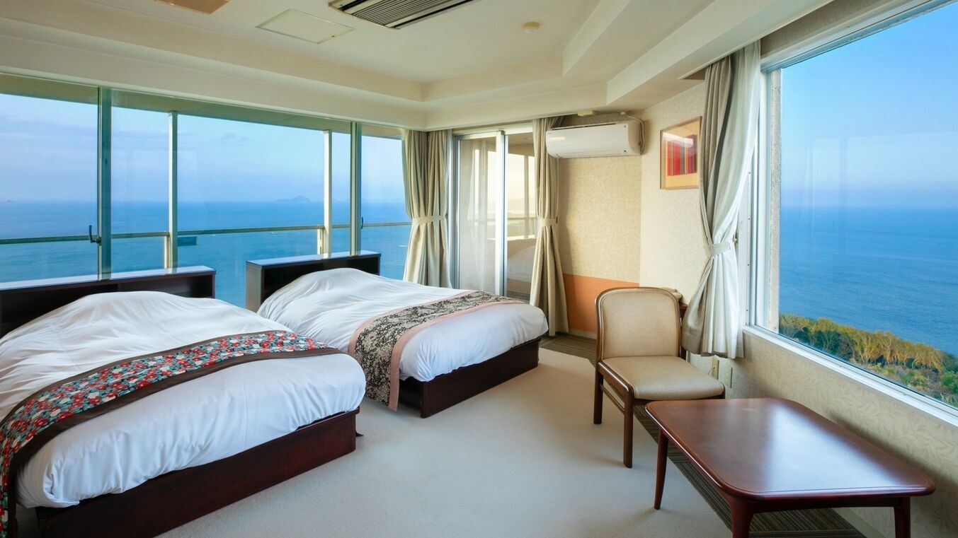 A hotel room with two beds, a chair, a table, and a large window with a view of the sea.