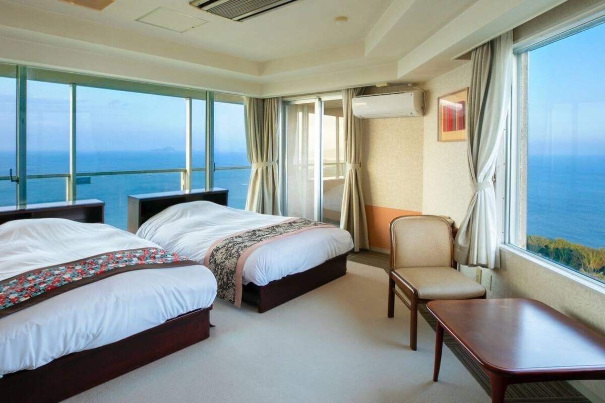 A hotel room with two beds, a chair, a table, and a large window with a view of the sea.