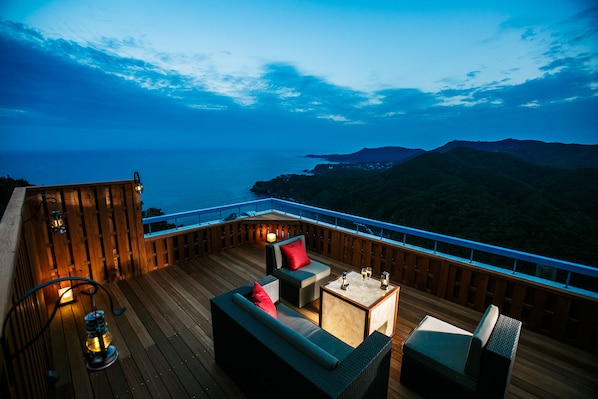 Una terraza de madera con mobiliario exterior, incluyendo un sofá y sillones, con vistas a un paisaje costero con colinas y el mar.