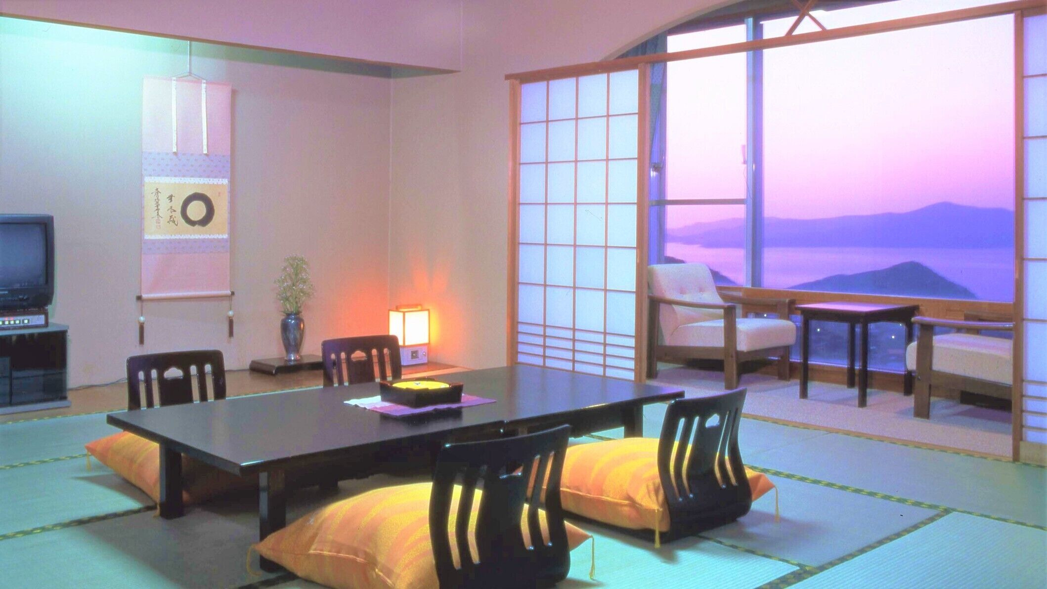 A traditional Japanese-style room with tatami flooring, a low table, chairs, a television, and a large window with a view.