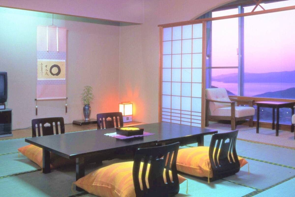 A traditional Japanese-style room with tatami flooring, a low table, chairs, a television, and a large window with a view.