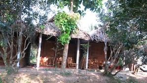 Une construction traditionnelle en bois, dotée d’un toit de chaume, entourée d’arbres et bordée d’un petit étang.