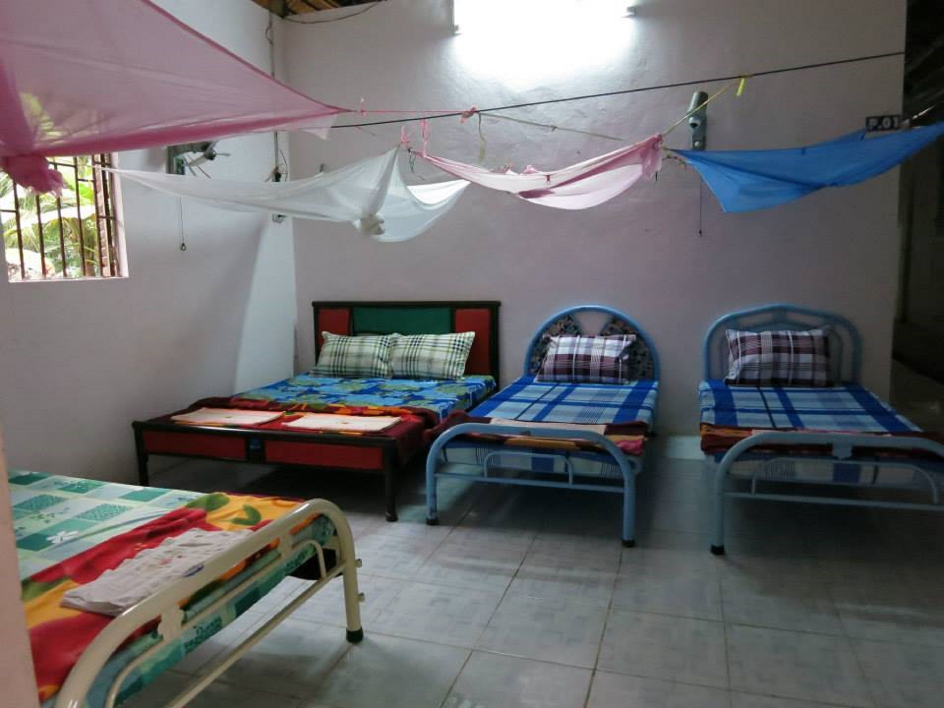 A room with three beds, each with a different headboard design. There is a window on the left side, and a mosquito net hanging above the beds.