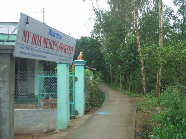 A sign welcoming visitors to My Hoa Mekong Homestay, indicating a rural setting with a pathway leading through a forested area. - My Hoa Mekong Homestay (Cai Von)