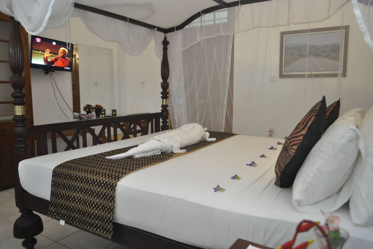 A neatly made bed with a white towel, a patterned blanket, and decorative pillows. A television is mounted on the wall, and a picture frame is visible.