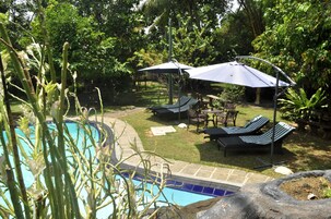 Un área de piscina con sillones reclinables y sombrillas, rodeada de exuberante vegetación.