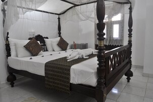 Una cama con dosel, ropa de cama blanca, almohadas estampadas y una mesita de noche con lámpara.