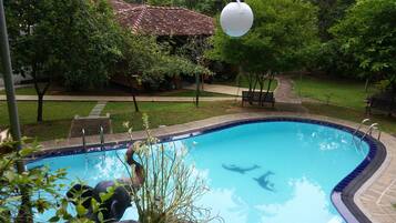 Una piscina con borde de azulejos, rodeada de vegetación y un pequeño edificio con techo de tejas.