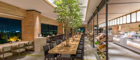 A long dining table with chairs, a brick wall, and a large tree in the center.