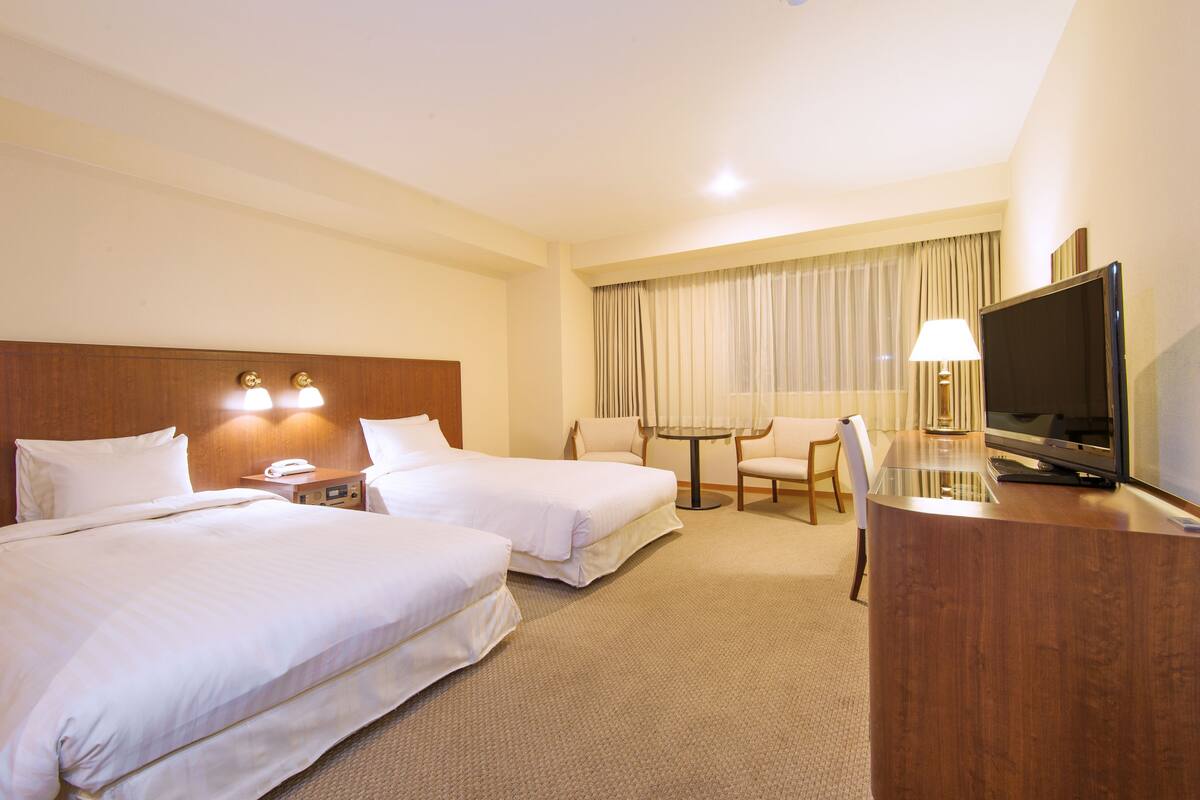 A hotel room with two beds, a desk with a television, a lamp, and a small table with chairs.