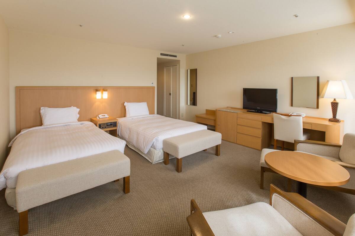 A hotel room with two beds, a desk, a chair, a television, and a lamp.