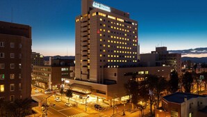 A modern Art Hotel with a prominent sign, surrounded by urban buildings and a well-lit street at dusk. - Art Hotel Asahikawa (Asahikawa)
