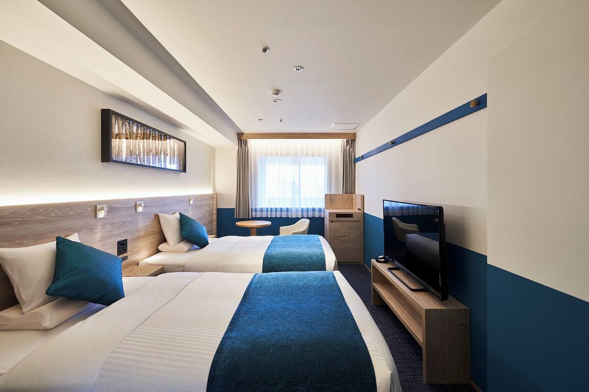 A hotel room with two beds, a TV, a desk, and a window with curtains.