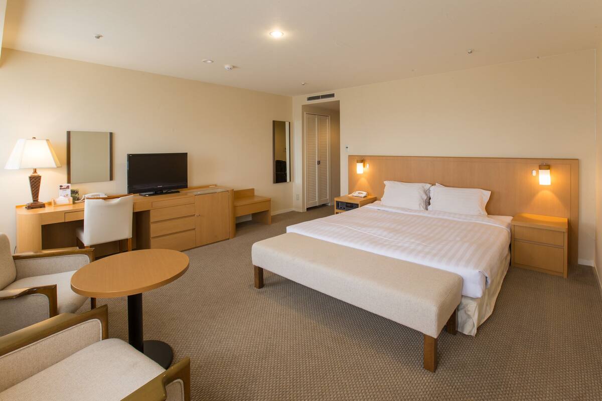 A hotel room with a bed, bedside tables, a desk with a television, a chair, and a small round table.