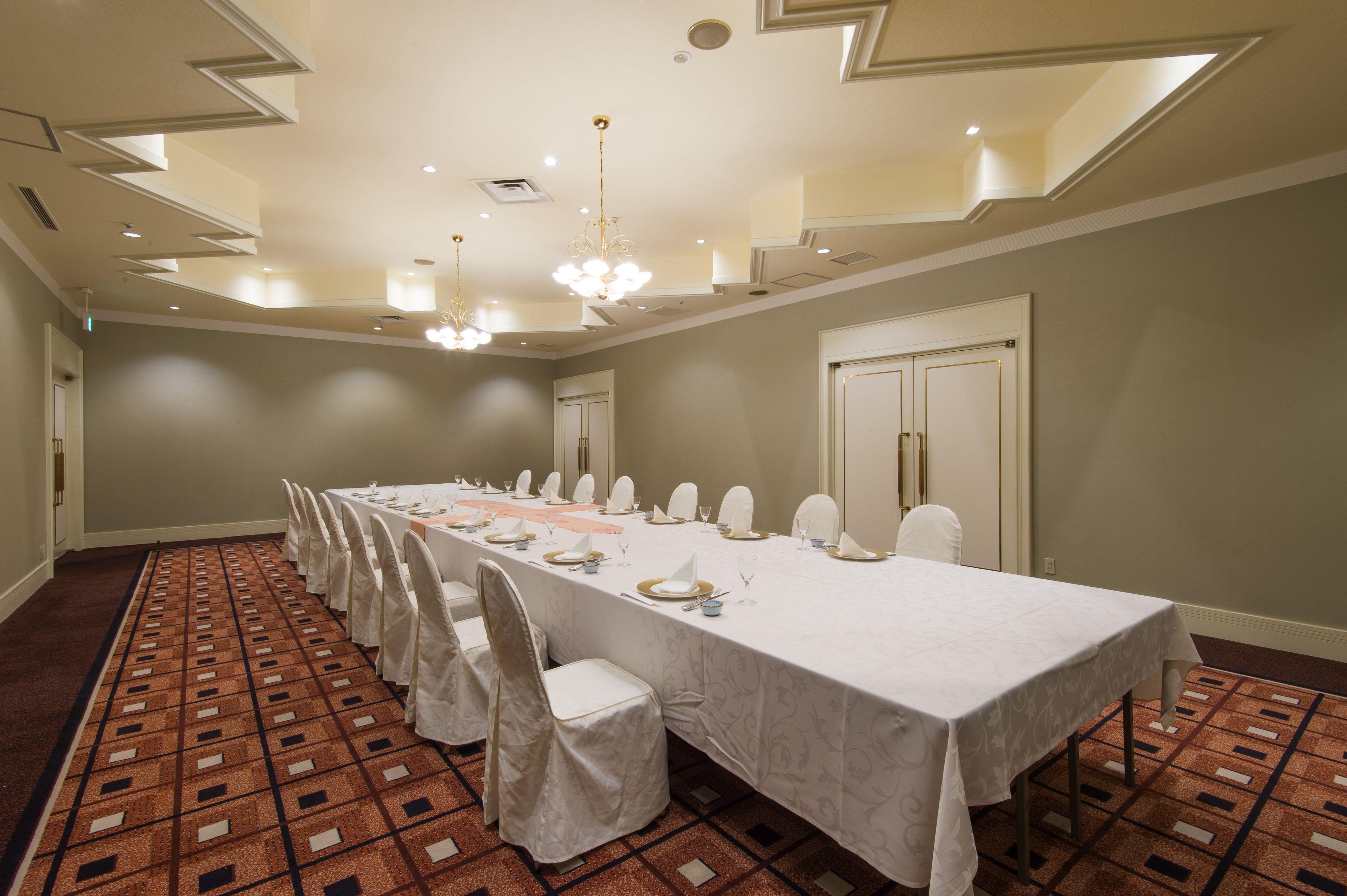Un salón de conferencias con una larga mesa dispuesta para una reunión, sillas blancas y una alfombra estampada.