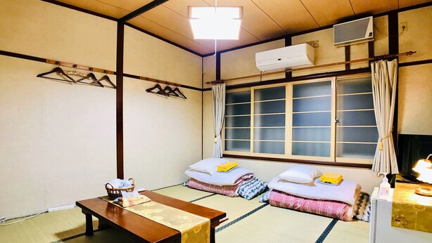 A traditional Japanese room with tatami flooring, a low wooden table, two futons with pillows, a hanging air conditioner, and a window with curtains. - Pension Puppy Tail (Hakodate)