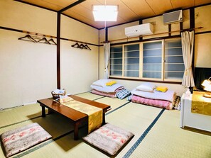 Una habitación japonesa tradicional con suelo de tatami, una mesa de madera baja, dos futones con almohadas, un aire acondicionado colgante y una ventana con cortinas.