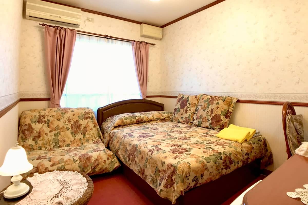 A bedroom with a bed, two chairs, a bedside table with a lamp, and a window with curtains.