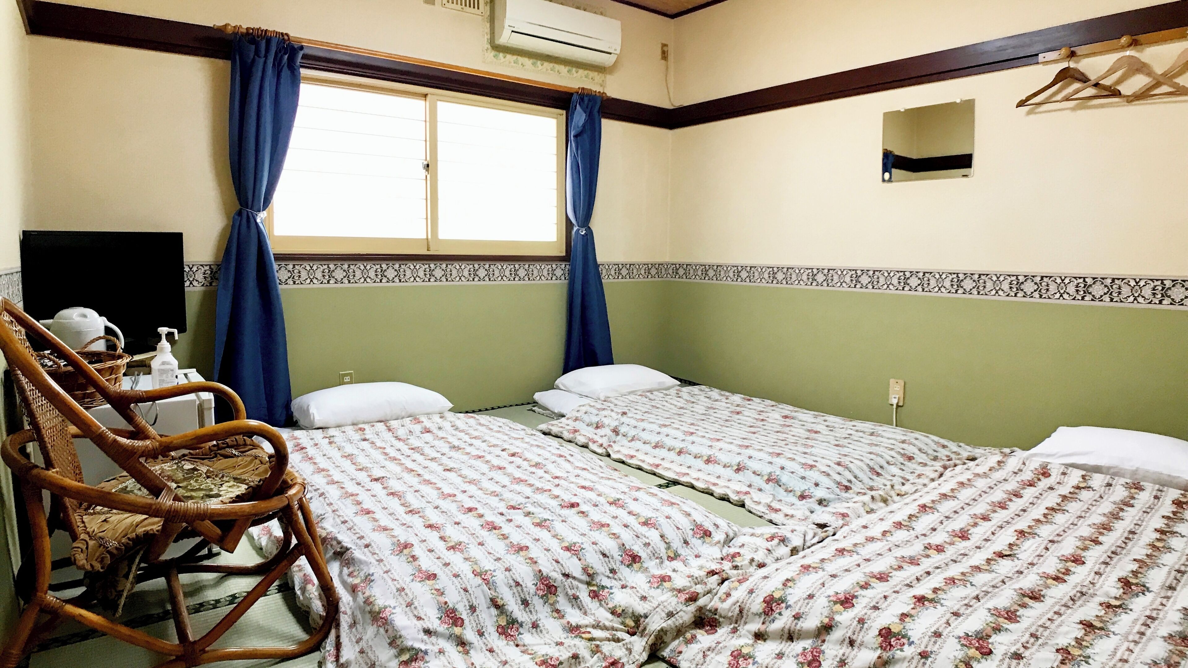 A room with two beds, a wooden rocking chair, a small table with a bottle, and a wall-mounted air conditioner.