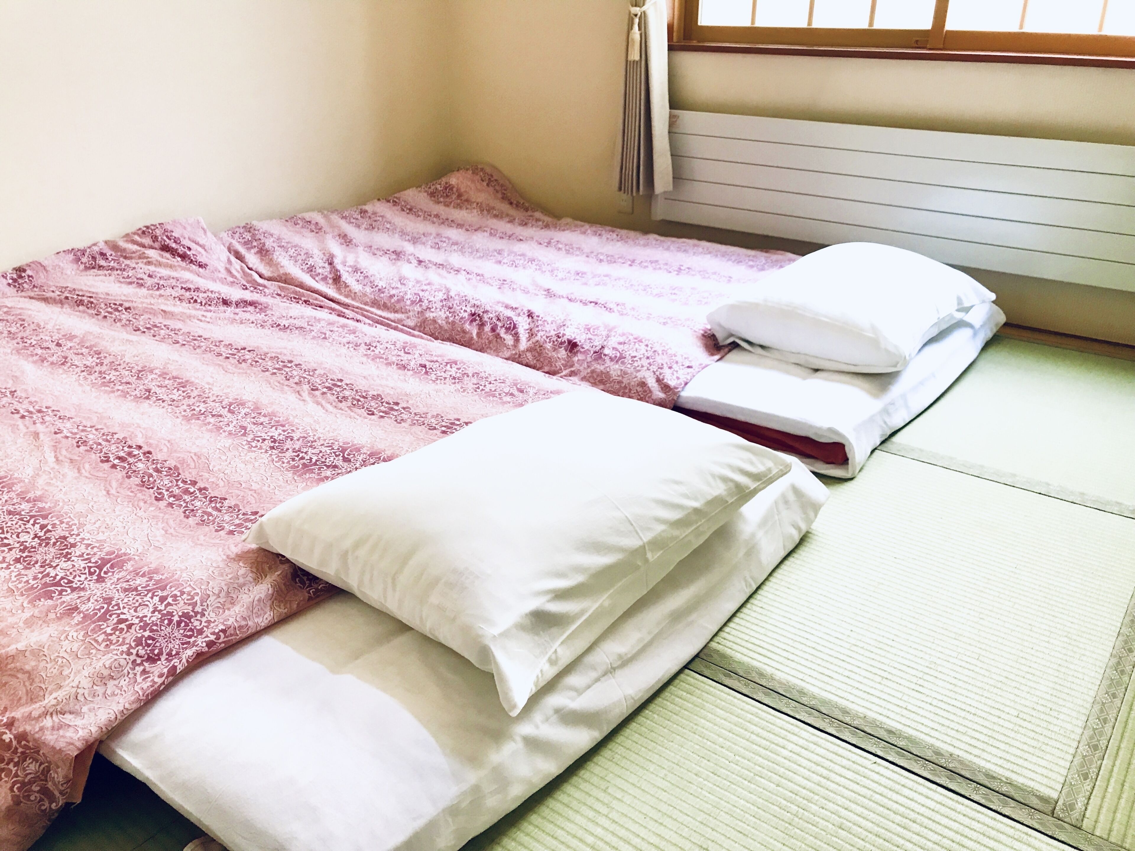 A single bed with a pink and white patterned bedspread and white pillows, a green mat, and a window with blinds.