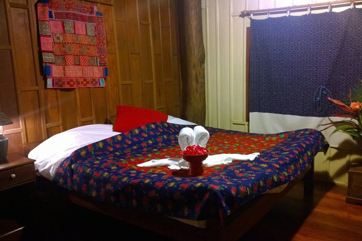 A neatly made bed with floral bedding, a red pillow, and a small red vase with flowers.