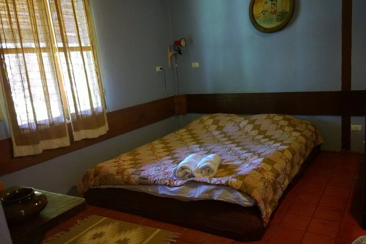 A small, cozy room with a single bed, a wooden headboard, a small table with a bowl, and a wall clock.