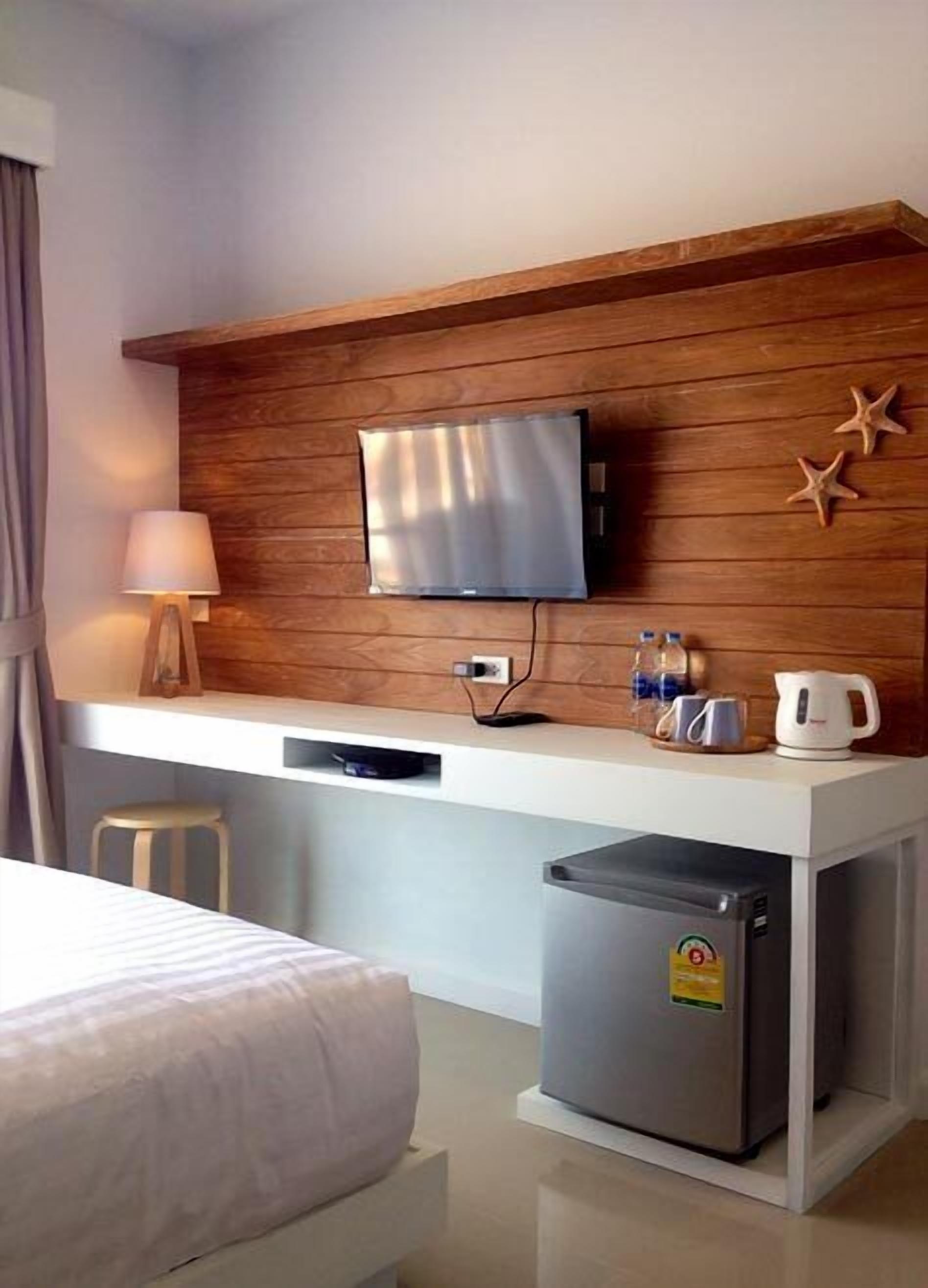 A hotel room with a wooden accent wall, a flat-screen TV mounted above a white desk, a mini-fridge, and a bed with a white comforter.
