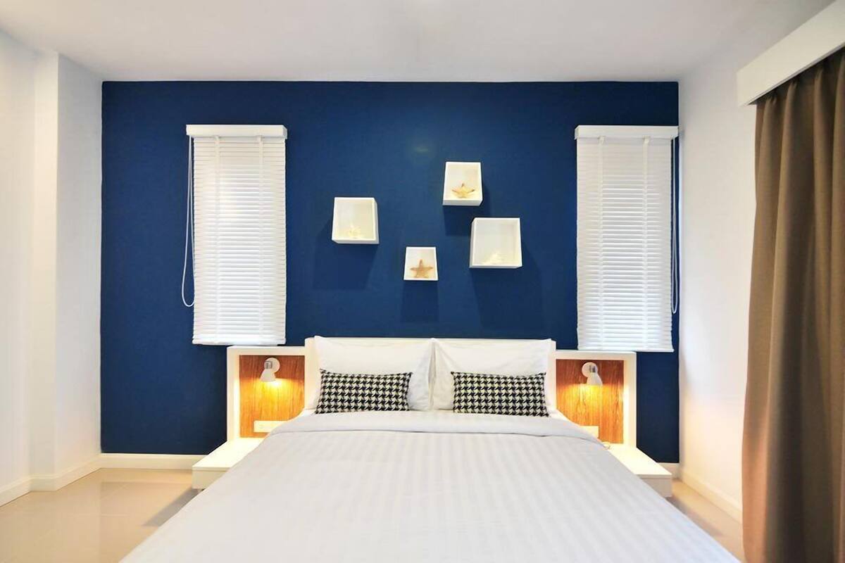 A bedroom with a large bed, two bedside tables with lamps, a blue accent wall, and two windows with white blinds.