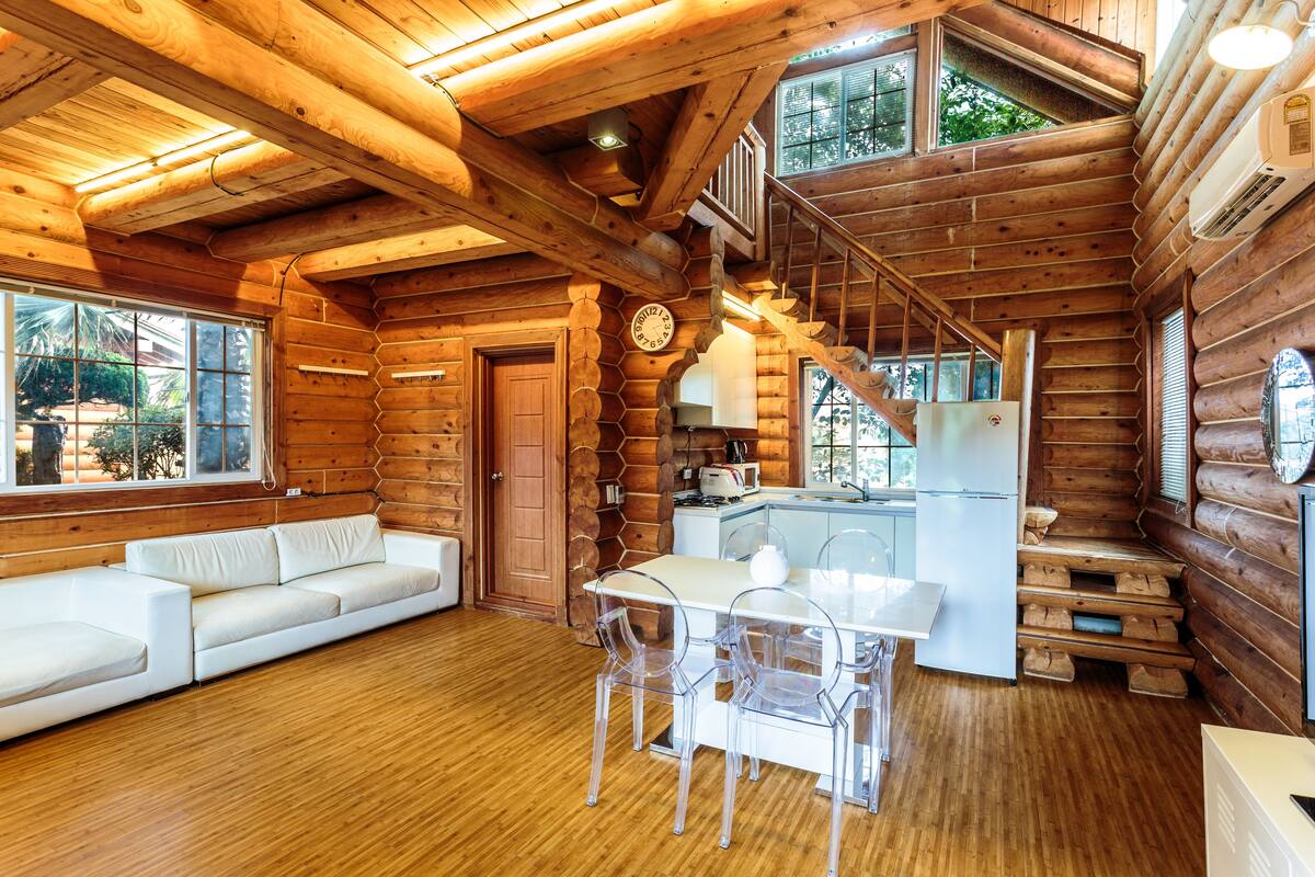 A log cabin interior with a wooden staircase, a white sofa, a dining table with chairs, and a kitchen area.