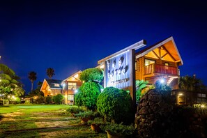 A well-lit, two-story wooden house with a large signboard, surrounded by neatly trimmed bushes and a stone pathway. - Ocean Log Pension (Seogwipo)